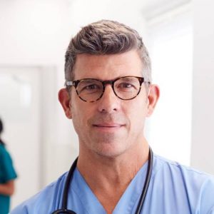 Portrait Of Mature Male Doctor Wearing Scrubs Standing In Busy Hospital Corridor